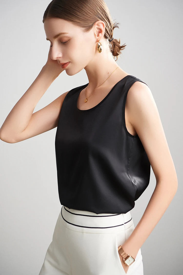 A woman wearing a black sleeveless silk top and white pants on a plain background