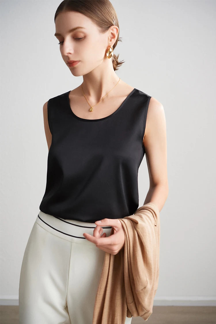 A woman wearing a black sleeveless silk top and white pants, holding a beige cardigan against a plain background.