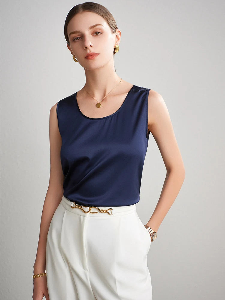 A woman wearing a navy blue sleeveless silktop and white pants against a gray background