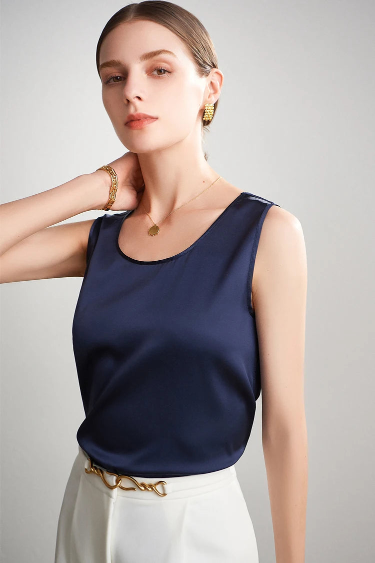 Woman wearing a navy blue sleeveless tsilk op with gold jewelry on a light gray background